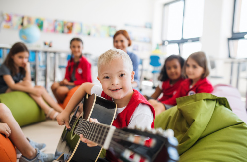 A group of children in a classroom, the child in centre focus has light blonde hair, appears to have previously had cleft lip & palate surgery, and is playing a guitar while looking confidently into the camera.
