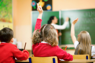 A pupil raises their hand in a classroom while the teacher writes on the board.