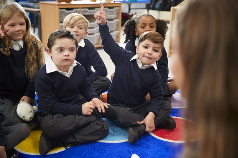 School children sat listening to a teacher, with one child with a raised hand
