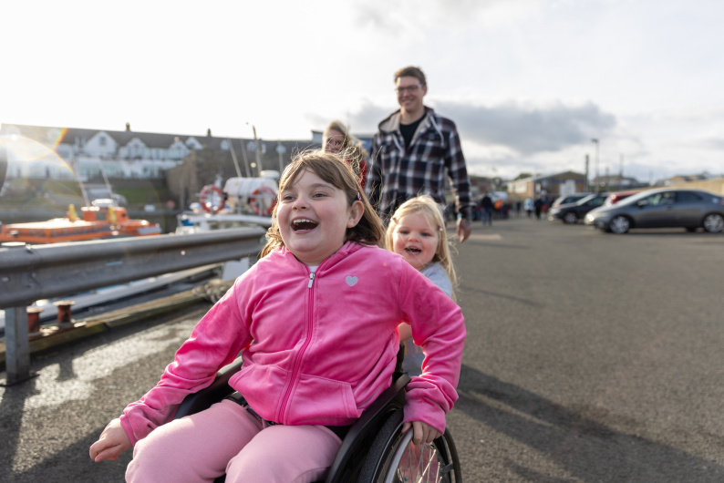 Girl in wheelchair smiling ahead of her little sister and parents